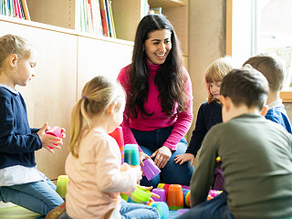 Eine Erzieherin sitzt in einer Krippe mit mehreren Krippenkindern im Kreis. Sie stapeln und sortieren Becher nach Farben.